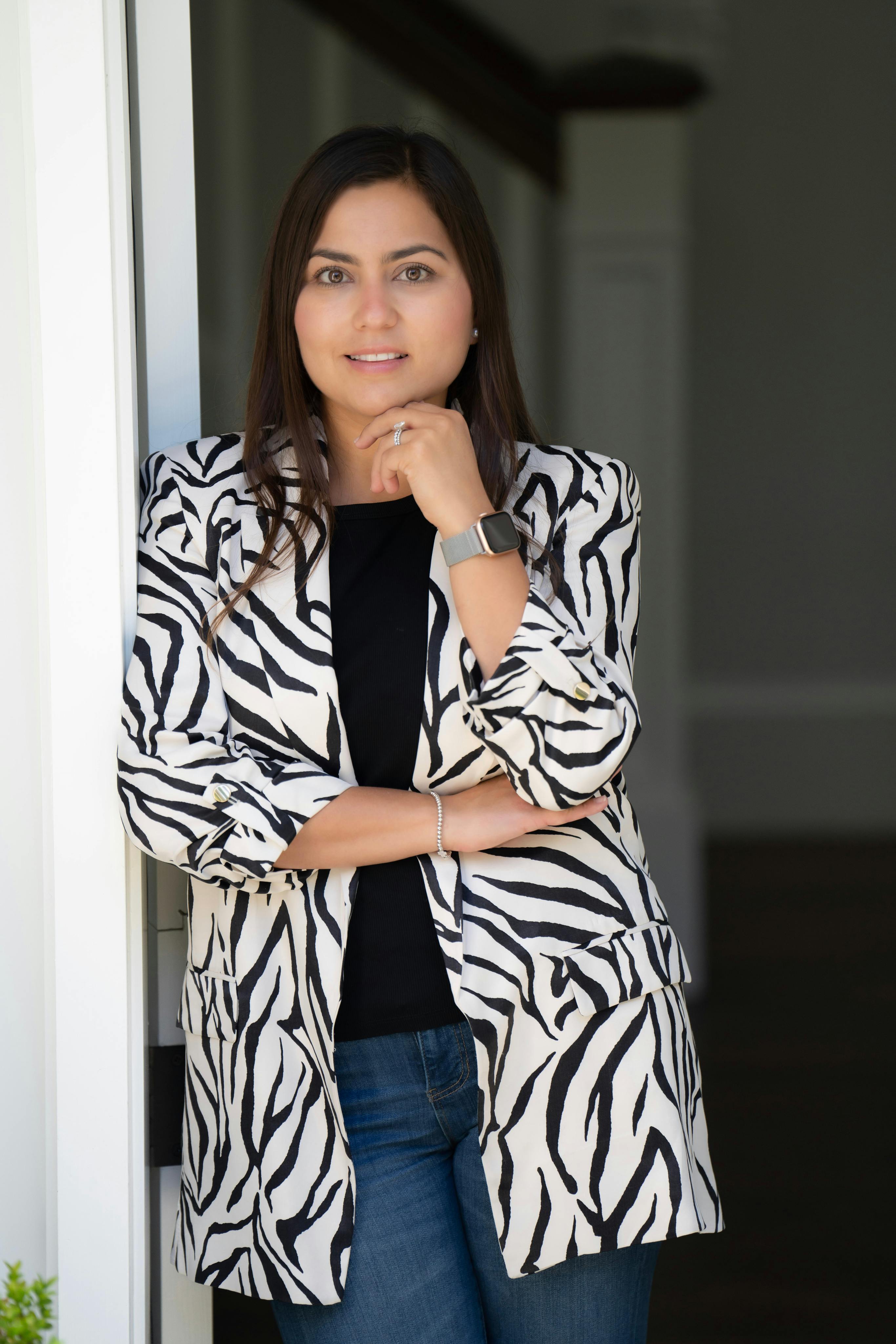 Andrea Brink stands leaning against a white doorframe. She is wearing a striking zebra print blazer over a black top, paired with blue jeans. Her arms are crossed, and she accessorizes with a silver watch and a simple bracelet on her left wrist. Her gaze is direct and thoughtful, with a slight smile on her face, suggesting a professional and approachable demeanor.