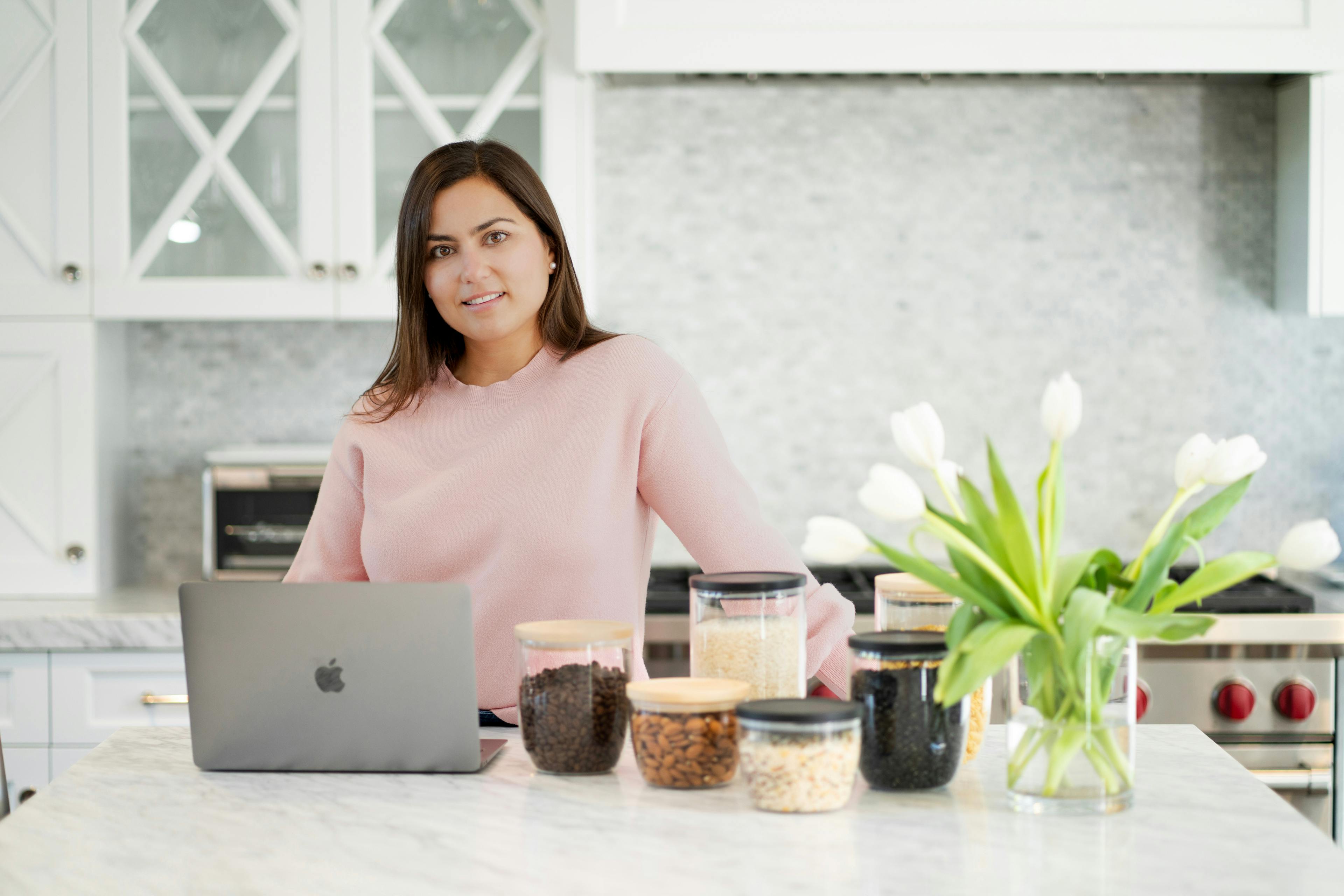 Andrea Brink wearing a soft pink sweater, stands behind a marble kitchen counter. She leans slightly towards an open laptop, showing an engaged expression. To her right, there are clear jars with various kitchen staples like rice, coffee beans, and almonds. A vase with white tulips adds a fresh touch to the serene kitchen setting, which features white cabinetry and a modern backsplash.