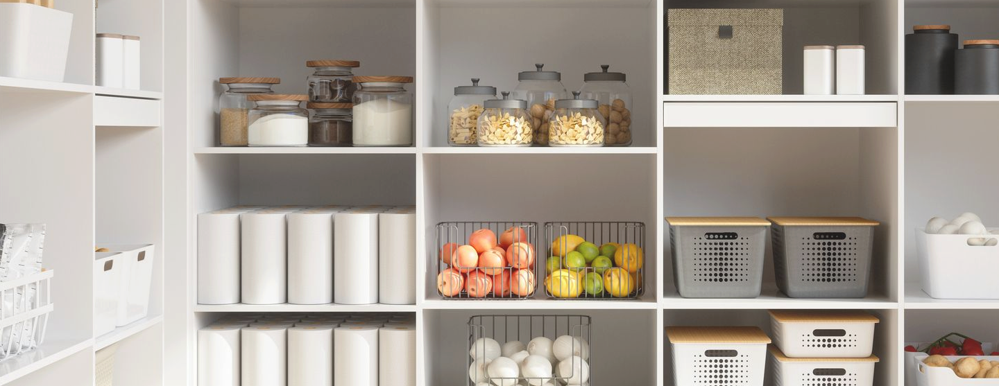 A well-organized pantry with an array of neatly arranged shelves against a light taupe wall. The shelves house a variety of food storage containers, glass jars filled with pasta, beans, and spices, and woven baskets containing kitchen towels. Lower shelves display a collection of fresh fruits in wire baskets, ceramic bowls with eggs, and several bottles of beverages. The clean and modern design is complemented by the warm wooden flooring.